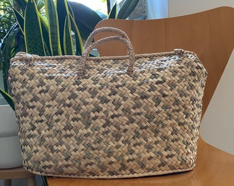 May include: A woven straw tote bag with a light brown and beige pattern. The bag has two handles and is sitting on a wooden chair.