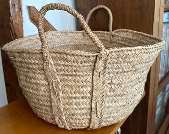 May include: A woven straw basket with two arched handles. The basket is light brown and has a wide, open design. The handles are tightly woven and appear sturdy. The basket sits on a wooden surface.
