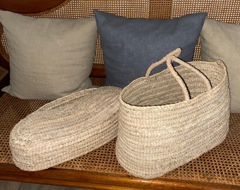 May include: Two woven straw baskets, one oval with handles and the other rectangular with a lid, sit on a wooden bench. The bench has a curved back and woven cane detailing. Behind the baskets are three linen pillows in neutral tones.
