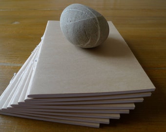 May include: A stack of blank, beige notebooks with a smooth, grey stone on top. The notebooks have white stitching along the spine.