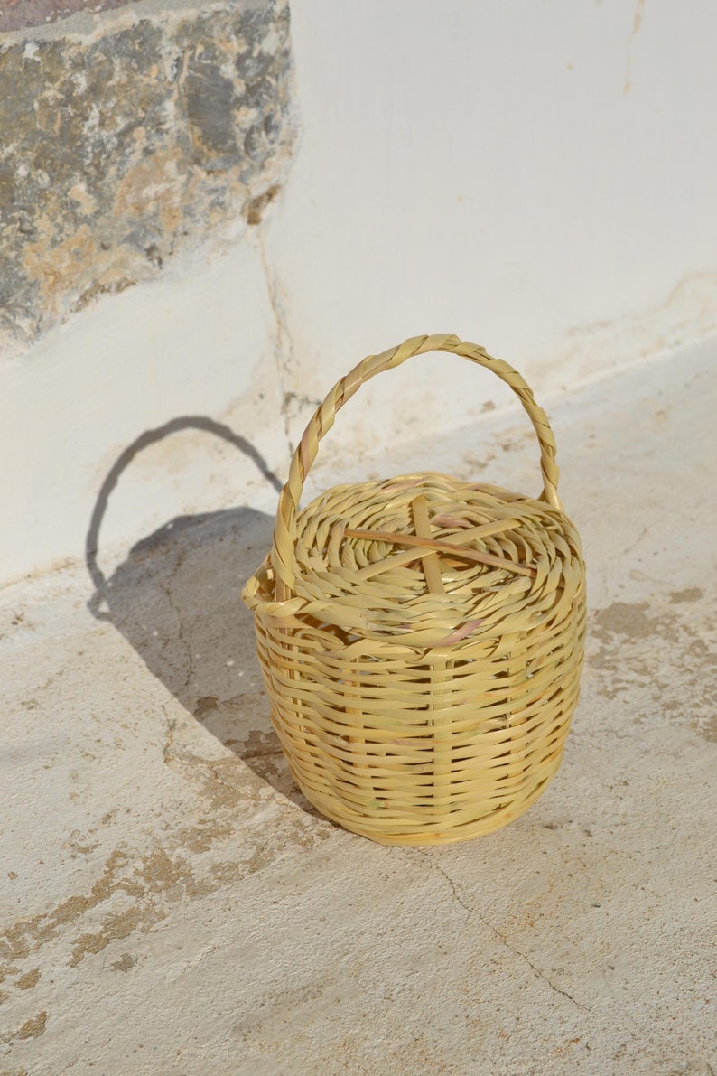 May include: A small, woven basket with a lid and a handle. The basket is made of natural materials and has a light brown color.