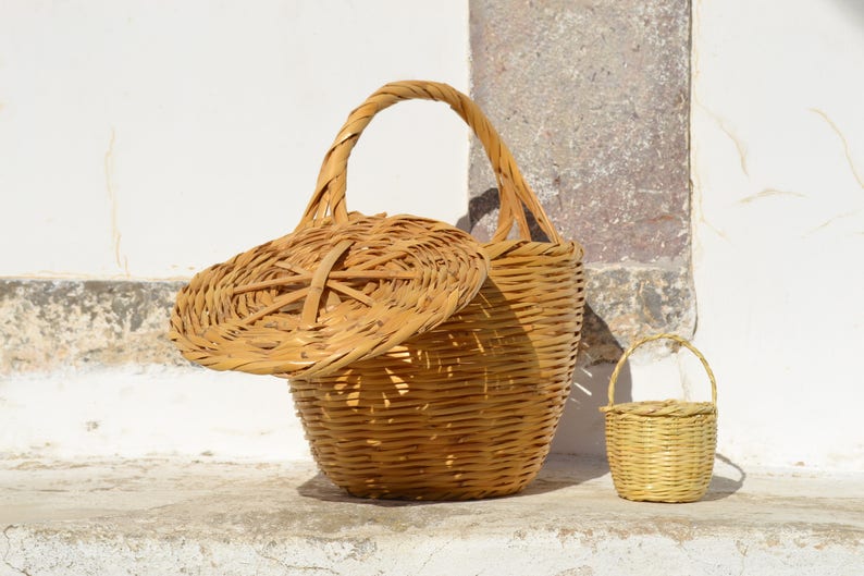 May include: Two woven wicker baskets, one large with a lid and a smaller one with a handle. The baskets are light brown and have a natural, rustic look.