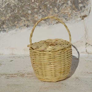 May include: A small, woven basket with a handle. The basket is made of natural materials and has a light brown color.