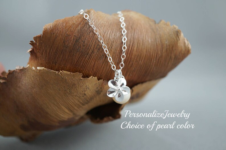 May include: A silver necklace with a flower pendant and a pearl. The pendant is made of silver and has a delicate flower design. The pearl is white and is set in the center of the flower. The necklace is on a brown wooden surface.  Personalize Jewelry Choice of Pearl color