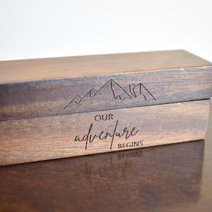 May include: A rectangular wooden box with a hinged lid. The box is stained in shades of brown and features a mountain range design and the words "Our adventure begins" engraved on the front.