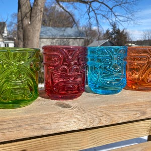 Op de afbeelding: Vier kleurrijke tiki-glazen in groen, rood, blauw en oranje staan op een houten oppervlak. Elk glas heeft een gedetailleerd tiki-ontwerp, met een cilindrische vorm en een textuur. De glazen zijn in een rij gerangschikt en tonen hun levendige kleuren.