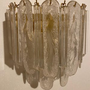May include: A clear glass wall sconce with a gold-colored metal frame. The sconce features vertical glass rods and frosted glass panels with a feather-like design. The fixture is designed to be mounted on a wall.