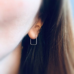 May include: A close-up of an ear with a small, square, silver earring. The earring has a minimalist design. Two gold hoop earrings are also visible. The background is blurred.