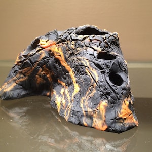 May include: A black and orange ceramic sculpture with a rough, textured surface. The sculpture has several holes and a unique, abstract shape.