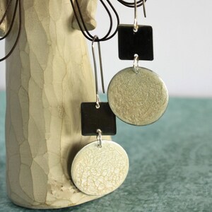 May include: Silver-tone dangle earrings with a geometric design. Each earring features a black square and a textured, light-colored circle. The earrings hang from silver hooks, creating a modern and stylish accessory.