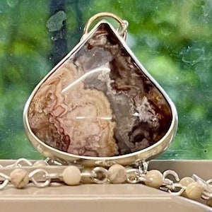 May include: A teardrop-shaped pendant with a silver-colored bezel. The pendant features a polished stone with a mix of brown, beige, and white patterns. A delicate beaded chain with small, round beads is attached to the pendant.