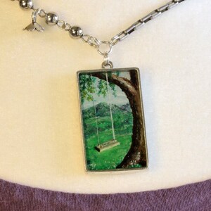 May include: A silver chain necklace with a rectangular pendant featuring a painted image of a swing hanging from a tree in a green field.