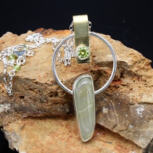 May include: A silver and gold pendant necklace with a green gemstone and a long, light green stone. The pendant is hanging from a silver chain.