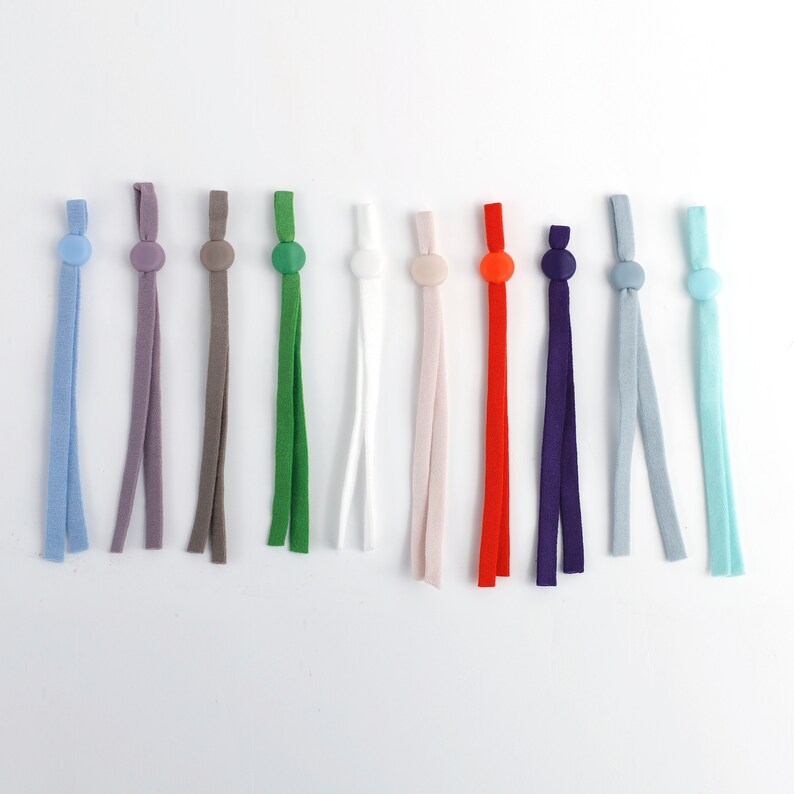 May include: Assortment of 12 colorful elastic mask strap extenders with button closures. The extenders are made of a soft, stretchy material and come in a variety of colors, including blue, gray, brown, green, white, pink, red, purple, light blue, and turquoise.