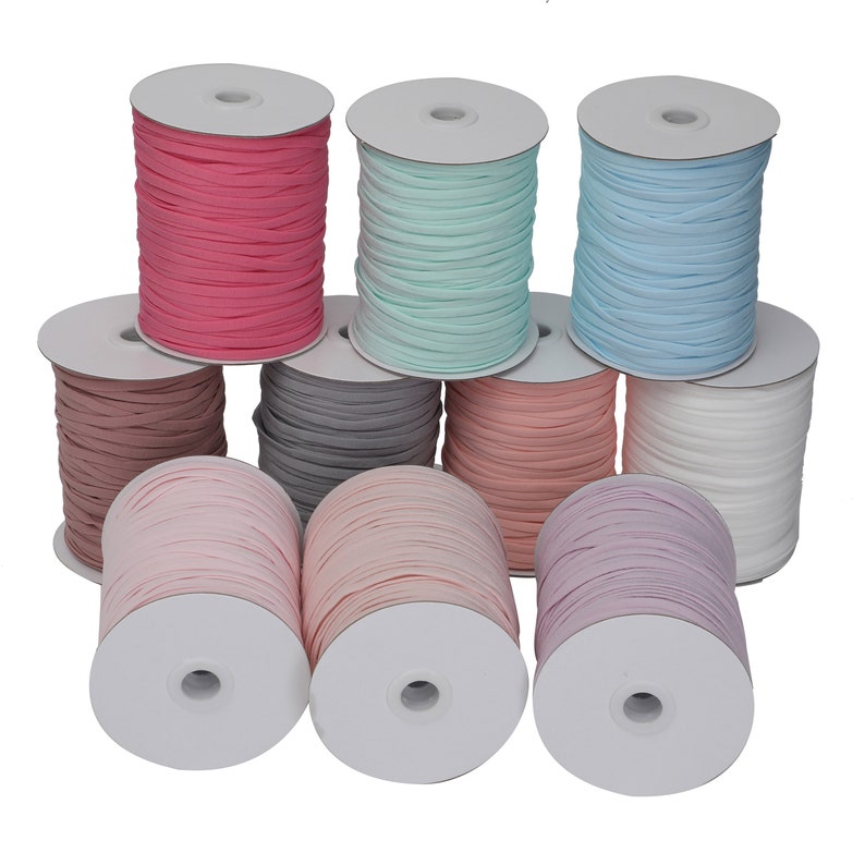 Op de afbeelding: Negen spoelen met gekleurde elastische koord, elk gewikkeld rond een witte kartonnen spoel. De kleuren zijn roze, lichtblauw, grijs, lichtroze, perzik, wit en donkerroze.