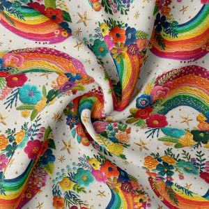 Puede incluir: Una tela blanca con un patrón repetido de arcoíris coloridos y ramos de flores. Los arcoíris están bordados con hilos de diferentes colores y las flores son en tonos de rosa, azul, amarillo y verde. El fondo es beige claro con estrellas doradas brillantes.