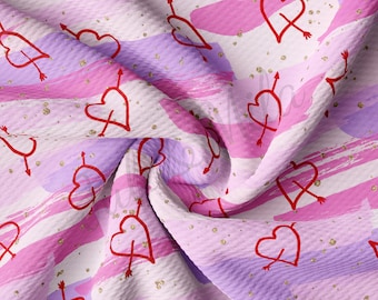 May include: Pink and purple fabric with a white background. The fabric has a textured waffle weave and is covered in a repeating pattern of red hearts with arrows through them. The hearts are outlined in gold glitter.