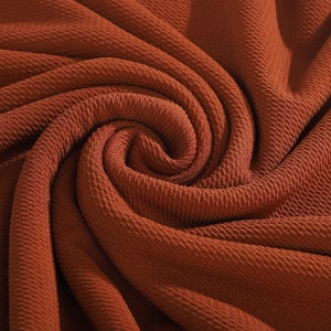 May include: Close-up of a rust-colored fabric with a textured, waffle-like pattern. The fabric is folded and draped, creating a swirling, abstract design.