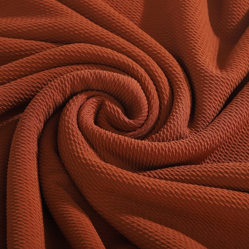 May include: Close-up of a rust-colored fabric with a textured, waffle-like pattern. The fabric is folded and draped, creating a swirling, abstract design.