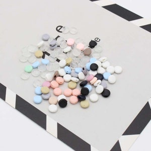 May include: Assortment of colorful plastic beads in various shades of blue, pink, beige, gray, and black. The beads are round and have a hole in the center. There are also clear plastic beads.