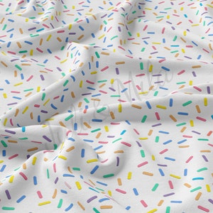 Op de afbeelding: Witte stof met een herhalend patroon van kleurrijke sprinkels. De sprinkels zijn in verschillende tinten blauw, groen, geel, oranje, roze en paars.