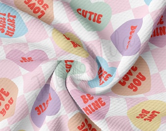 May include: A white and pink checkered fabric with a repeating pattern of colorful heart-shaped candy with various messages, including "Be Mine", "Bug Love", "Cutie", "Love You", and "Sweet Heart".