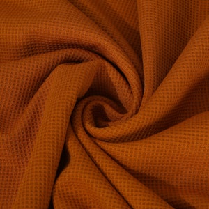 May include: A close-up of a rust-colored waffle knit fabric. The fabric is folded and draped, showing the texture of the knit.
