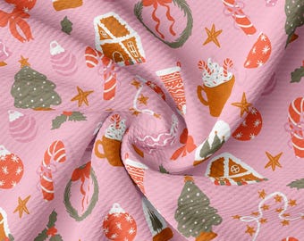 Peut inclure: Tissu rose avec un motif répété d'illustrations sur le thème de Noël. Le motif comprend des maisons en pain d'épices, des cannes en sucre, des sapins de Noël, des boules et des tasses de chocolat chaud.