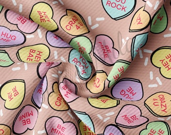 May include: A pink fabric with a repeating pattern of colorful candy hearts. The hearts have various messages on them, including "Hey Bae", "Be Mine", "Crazy 4 U", "You Rock", and "Hug Me".