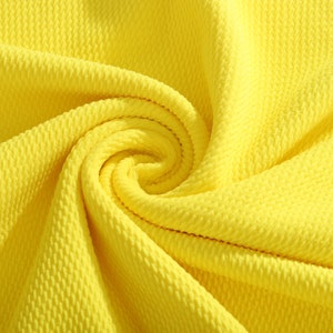May include: Close-up of a bright yellow fabric with a textured, waffle-like pattern. The fabric is folded and draped, showing the texture and the soft, pliable nature of the material.
