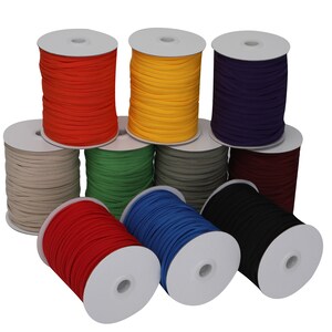 Op de afbeelding: Spoelen met gekleurde elastische koord in verschillende kleuren, waaronder rood, geel, groen, blauw, zwart, wit, paars en bruin. De spoelen zijn in een cirkel op een witte achtergrond gerangschikt.