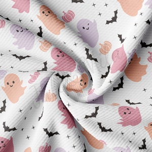 May include: White fabric with a repeating Halloween pattern. The design features smiling ghosts in shades of pink, peach, and lavender, along with black bats, orange pumpkins, and small black crosses. The fabric has a textured appearance.