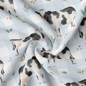 May include: A blue and white gingham fabric with a repeating pattern of black and white cows and green leaves. The fabric is soft and textured.