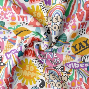 May include: A white fabric with a colorful pattern of various words and symbols. The words include "So Happy", "Feel Good", "You've Got This", "Yay", "Vibes", and "Do It!" The symbols include a rainbow, a sun, a pineapple, and a yin-yang symbol.