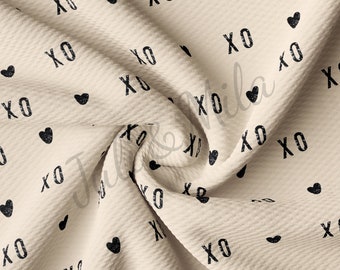 May include: Cream-colored fabric with a repeating pattern of black "XO" text and heart shapes. The fabric has a textured surface, and the design is evenly distributed across the material. The overall aesthetic is simple and romantic.