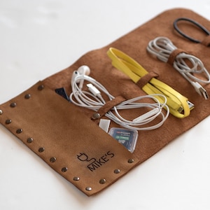 Peut inclure: Un étui enroulable en cuir marron avec une fermeture à glissière et une inscription personnalisée qui dit "Mike's". L'étui est conçu pour contenir et organiser divers cordons et câbles, y compris un câble de charge jaune, un cordon de casque blanc et un câble USB noir.