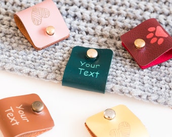 May include: Five folded leather labels with metal rivets in pink, dark red, dark green, tan, and yellow. The labels feature engraved designs including a heart, a paw print, and the text Your Text, attached to a grey knitted fabric surface.
