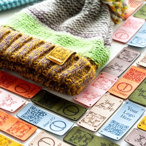 Puede incluir: Una colección de etiquetas de cuero de colores con un código QR y el texto "Your Logo Here" impreso en ellas. Las etiquetas están dispuestas sobre una superficie blanca junto a un gorro de punto con un patrón de rayas verdes, grises y amarillas.
