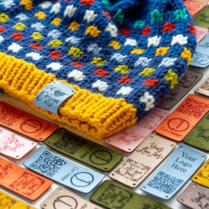 May include: A blue and yellow knitted hat with a colorful pattern of stars and dots. The hat is lying on a table with several leather tags with different designs and text, including "Your Logo Here" and "Out Design".