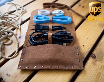 May include: Brown leather cord organizer with a flap closure and snap button. The organizer has multiple slots for cords and cables. The organizer is on a wooden surface. The text "UPS Express" is visible in the background.