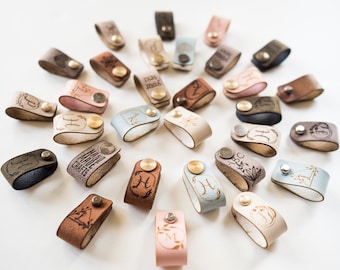 May include: A collection of leather cord keepers with various designs and colors. The cord keepers feature engraved initials, words, and symbols. The cord keepers are made of leather and have a brass button.
