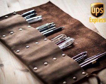 May include: Brown leather roll-up case with metal studs, holding a variety of pens and pencils. The case is rolled up and secured with a strap. The case is on a wooden surface.