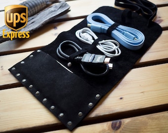 Op de afbeelding: Een zwarte leren kabelorganizer met zilveren studs, die verschillende kabels bevat, waaronder een blauwe HDMI-kabel, een witte USB-kabel en een zwarte kabel. De organizer is opgerold en heeft een band om hem gesloten te houden.