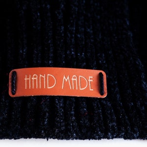 May include: A close-up of a black fabric with a brown leather tag that says "HAND MADE" in white lettering.