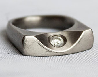 May include: A silver ring with a square-shaped band and a single, clear diamond set in the center.
