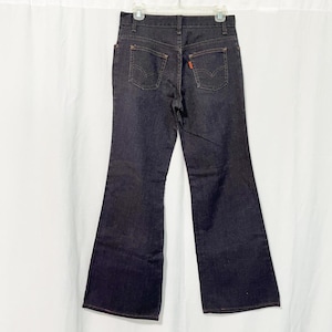 May include: Dark wash denim jeans with a flared leg. The jeans feature classic five-pocket styling, orange stitching, and a red logo tag on the back pocket. The jeans are hanging on a white hanger against a white backdrop.