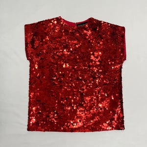 May include: A red sequined top with short sleeves. The top is covered in small, shiny sequins, creating a shimmering effect. The neckline and inner sleeves are a darker red. The tag reads "Schroeder's".