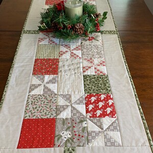 K&ouml;nnte beinhalten: Ein wei&szlig;er und gr&uuml;ner Patchwork-Tischl&auml;ufer mit einem roten und wei&szlig;en Sternenmuster. Der L&auml;ufer ist mit einem gr&uuml;nen Kranz und einer Kerze verziert.