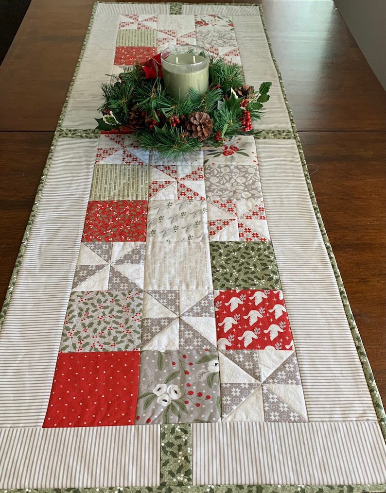 K&ouml;nnte beinhalten: Ein wei&szlig;er und gr&uuml;ner Patchwork-Tischl&auml;ufer mit einem roten und wei&szlig;en Sternenmuster. Der L&auml;ufer ist mit einem gr&uuml;nen Kranz und einer Kerze verziert.