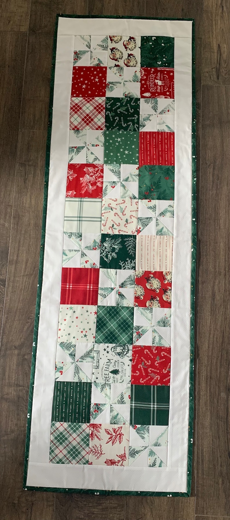 K&ouml;nnte beinhalten: Ein wei&szlig;er Tischl&auml;ufer mit gr&uuml;nem Rand zeigt ein Patchwork-Design aus roten, gr&uuml;nen und wei&szlig;en Stoffquadraten mit verschiedenen weihnachtlichen Mustern. Die Stoffquadrate sind in einem Raster angeordnet und zeigen Designs wie Zuckerstangen, Schneeflocken und Weihnachtsb&auml;ume.